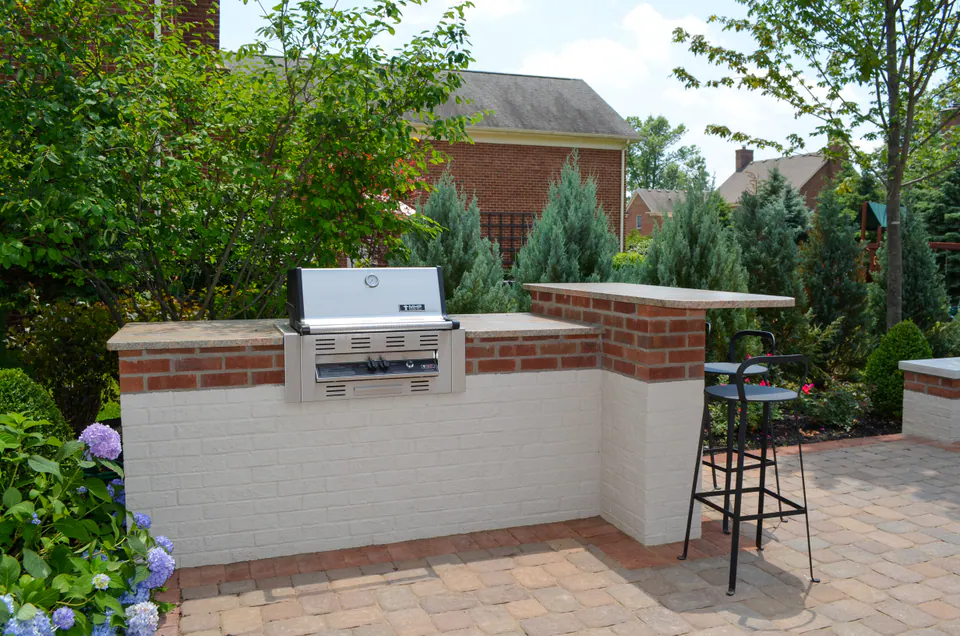 Painted brick outdoor grill counter with stainless grill, stone countertop, paver patio, hydrangeas, bar seating, and evergreen screen